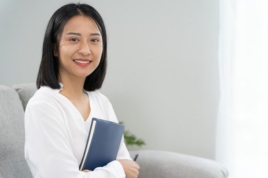 asian businesswoman smiling and holding a book in office. beautiful and good looking asian woman sits on the sofa. female portraits. 