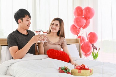 lovely couple and valentine's day concept. smiling asian man and woman drinking sparkling wine in bed at bedroom with red balloon and beautiful gift box.