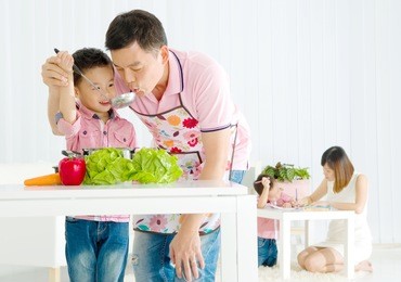 asian father and elder son are cooking in the kitchen while mother is teaching younger son doing schoolwork.