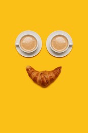 two cups of coffee with foam in white mugs cups with a breakfast morning croissant forming a smiling happy face on a yellow background with copy space and room for text