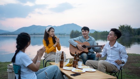 happy group of asian friends play guitar and sing enjoying camping and drinking beer