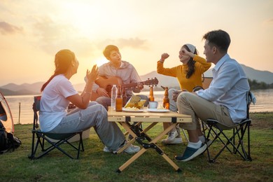happy group of asian friends play guitar and sing enjoying camping and drinking beer