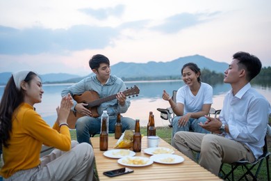 happy group of asian friends play guitar and sing enjoying camping and drinking beer