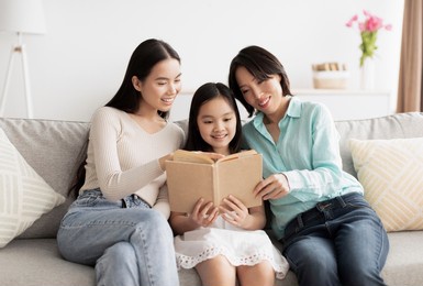 cute little asian girl reading book with her mommy and granny on couch at home. affectionate multi generation family enjoying interesting story together in living room