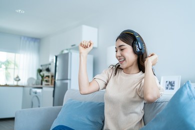asian girl listen to music and dance while sit on sofa in living room. attractive beautiful young woman put on headphone feeling happy to spend leisure time at home and enjoy weekend activity at home.
