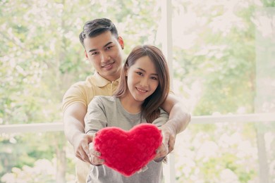 happy couple sitting on the sofa and being a man embracing his girlfriend with love in the living room and smile and holding a red heart. concept of romantic on valentine day. proposal and marriage