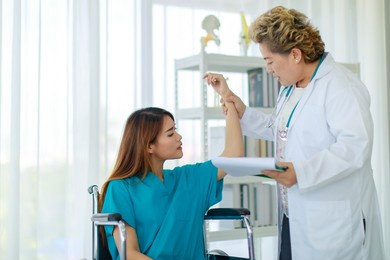 old senior professional female doctor in white lab coat standing using stethoscope listening to heartbeat of disabled handicapped woman patient sitting on wheelchair in hospital checkup ward room.