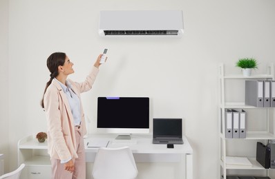 happy office worker or business lady turns on ac. smiling young woman in suit standing in office interior and switching wall air conditioner with remote to enjoy cool breeze on hot summer day at work
