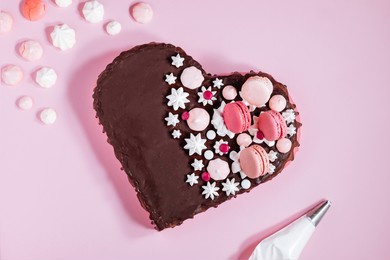 top view of pink heart shaped cake with chocolate glaze, meringues and macaroons on top as decoration. placed on pink background. valentine's day, mothers day, birthday, celebration concept.