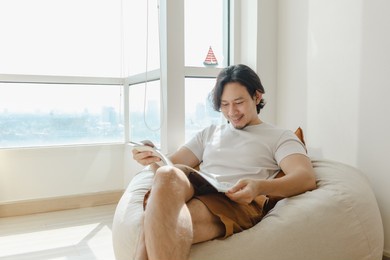 asian man relax and reading book on bean bag sofa in his apartment.