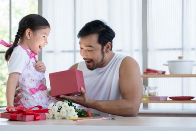 asian young healthy dad with beard and little mixed race daughter opening gift box with exciting or surprising face. family sitting at home kitchen celebrating father day or birthday special occasion