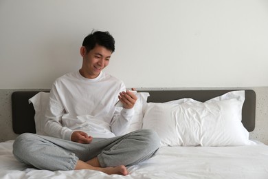 smiling asian young man sitting on bed, using and looking at phone at home