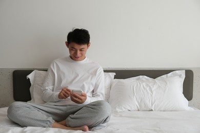 smiling asian young man sitting on bed, using and looking at phone at home