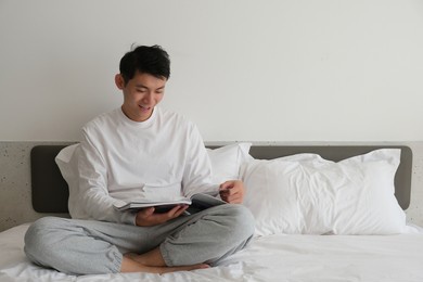 smiling asian young man sitting on bed reading a book