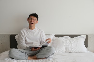 smiling asian young man sitting on bed reading a book, looking at camera