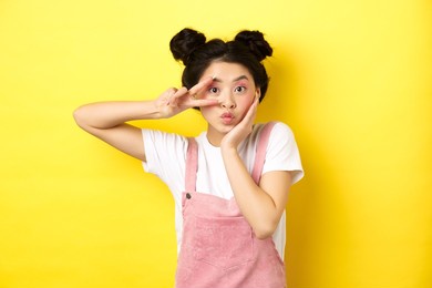 kawaii asian girl showing v-sign and pouting cute, making silly face with makeup, standing on yellow background