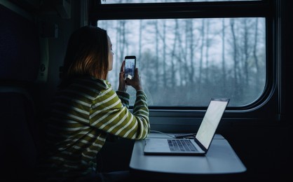 stylish woman sitting at a table in a train with a laptop and takes photos of evening views from the window of the compartment.