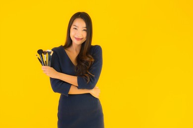 portrait beautiful young asian woman with makeup brush on yellow isolated background