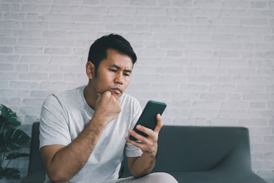 depression concept. asian man sad and checking mobile phone sitting on the sofa in the living room at home, asian man serious and worry while using mobile phone.
