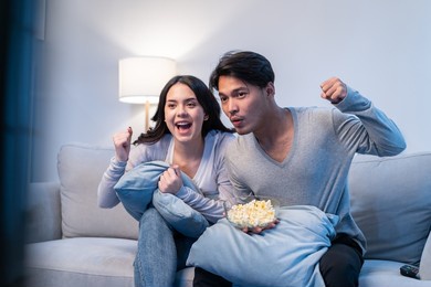 asian new marriage couple watching football sport game on television. excited man and woman fans sit on sofa in living room and watch tv sport game then celebrate team victory win in match at home.