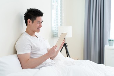 asian handsome man read online news from tablet in morning at home. happy attractive male lying down on bed in bedroom enjoy holiday weekend, using smartphone communication with technology in house.