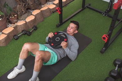 a fit asian man does weighted abdominal crunches with a plate while lying on a black mat. working out his core at his home gym.