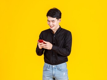 studio shot of millennial asian excited male businessman model in casual black long sleeve shirt and jeans standing holding red smartphone in hands playing virtual video game on yellow background.