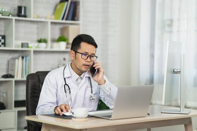 asian doctor man calling mpbile phone and using laptop to give consult to the patient via video call in the home office. telemedine and healthcare concept
