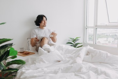 asian man reading book on the bed with breads and coffee in his apartment.