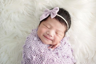 smiling newborn wrapped with purple swaddling and purple headband sleeping on comfortable white fur.              