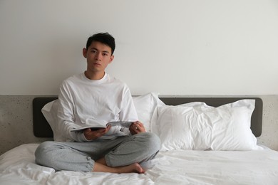 handsome asian young man sitting on bed holding a book, looking at camera