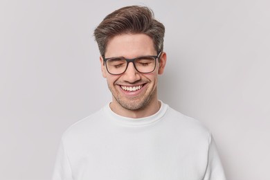photo of handsome cheerful man with dark hair smiles gladfully with closed eyes expresses positive emotions daydreams about something wears spectacles and casual jumper isolated on whie wall