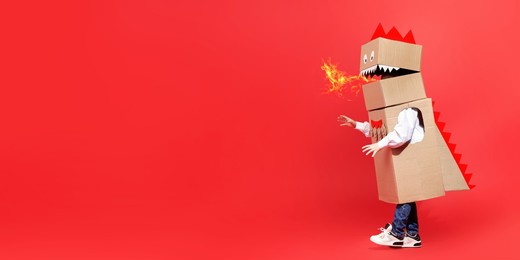 childhood dreams. funny little boy plays a cardboard dragon breathing fire and walking on a red background. fantasy, imagination. full-length studio portrait. copy space.