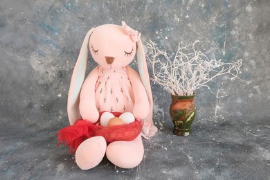a pink sleeping hare, a rabbit girl, a toy with long ears, sits, holding a basket with white chicken eggs in her paws. gray grunge background. easter concept, congratulations, customs. place for text