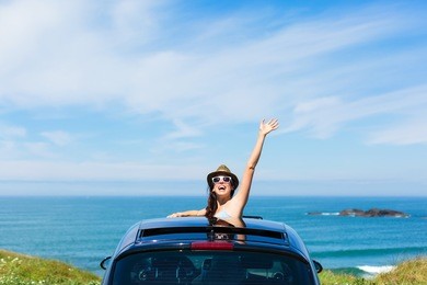 joyful woman waving on summer car travel vacation to the coast. brunette girl having fun leaning out vehicle sunroof towards the sea.