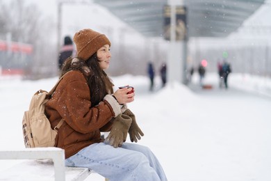 smiling asian girl in warm cozy winter clothes sitting on snow-covered railway platform with mug of hot tea in frozen hands, tired but happy traveler waiting for train to arrive, blurred background