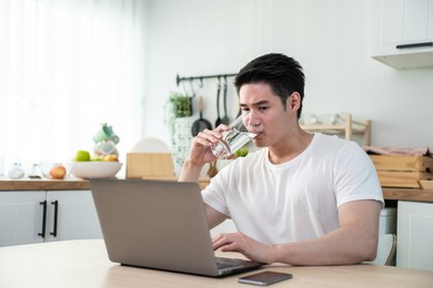 asian business man drinking miniral water while sit working from home. attractive handsome male feel happy, use laptop computer study learn online and sipping clean water for health care in kitchen.