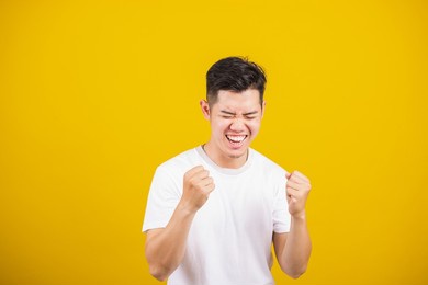 asian handsome young man smile positive shaking hands enthusiastic shouting yes for win competition, male raising his fists yes! with smiling delighted face, studio shot isolated on yellow background