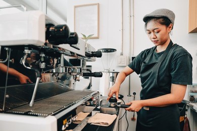 female barista presses ground coffee into portafilter using tamper. young asian coffeemaker woman standing at coffee machine in cafe preparing fresh hot espresso in the morning. small business owner.