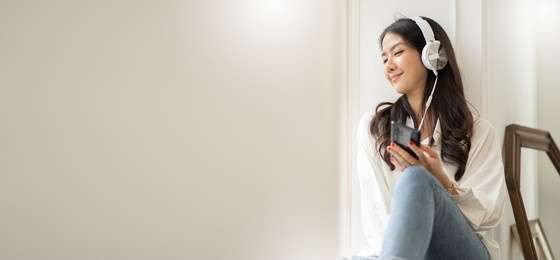 asian woman sitting at the window at home listening to music with headphones.beautiful young female at home using smartphone enjoys listening online music through headphones.music streaming concept.