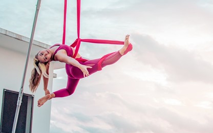 beautiful healthy strong woman wearing sport bra, relaxing, exercise yoga fly in air, aerial hoop on outdoor rooftop in morning. there are background of sky, sunlight flare, copy space advertisement