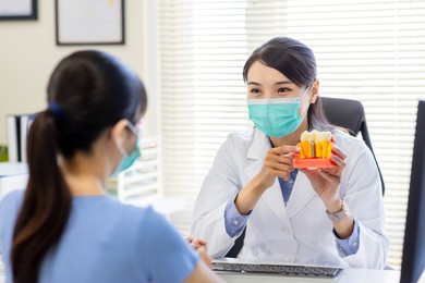 close up of asian female dentist introduces patient for dental implant by denture model while they wear protective face mask to prevent covid19