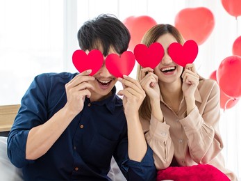 playful asian man and woman lovers  smiling and holding paper hearts near eyes during saint valentines day celebration. happy couple love concept.