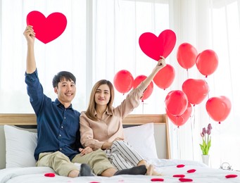 optimistic asian man and woman couple lovers looking at camera with smile and raising arms with paper hearts while resting on bed on saint valentine day