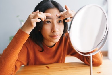 asian young woman squeezing acne skin and looking on the mirror at home, close- up of young women face