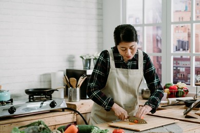 woman cutting carrot on wooden island table in modern kitchen at home. smiling asian chinese lady watching tv show on digital tablet and cooking dinner preparing healthy meal with fresh vegetables.