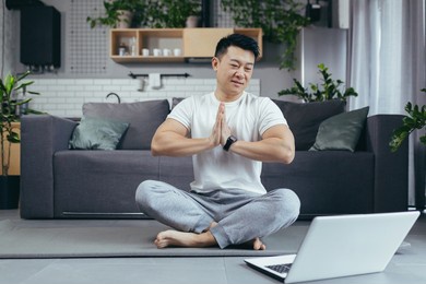 happy asian practices yoga at home, sitting in lotus position, man uses laptop for online classes, leads an active lifestyle