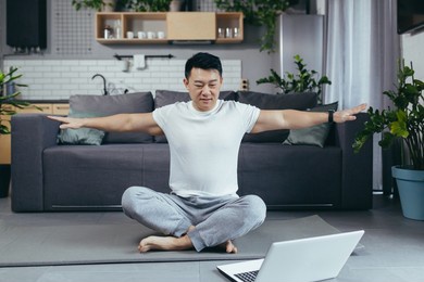 happy asian practices yoga at home, sitting in lotus position, man uses laptop for online classes, leads an active lifestyle