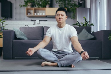 asian himself meditates at home in the morning, sitting on a sports mat, near the sofa in the living room, his morning, calm outside the lotus