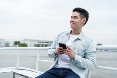 asian handsome male sit on ferry travel in the city for holiday trip. attactive young man backpacker tourist use mobile phone chatting message while sit on boat enjoy and happy spend time on vacation.